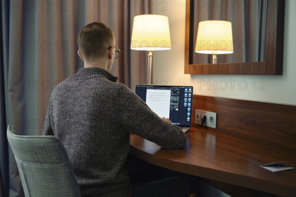 A person sits at a wooden desk in a hotel room, focused on his laptop screen. Soft lamp light