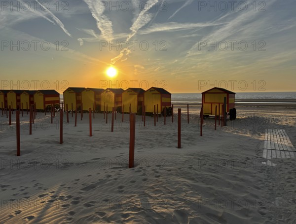 Beach huts at sunset with sand and sea in the background. The sky is streaked with clouds, De