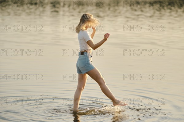 A woman walks through shallow water, splashing lightly as golden sunlight reflects off the surface.