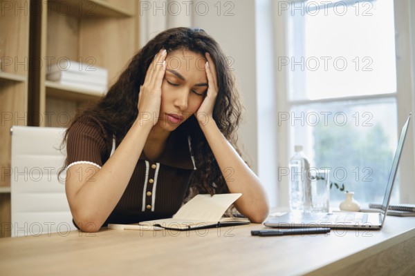 Tired young woman sits at her desk, visibly stressed and holding her head in her hands. The