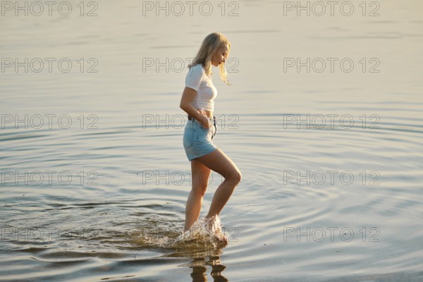 A young woman wearing a white t-shirt and denim shorts walks slowly through the shallow waters of a