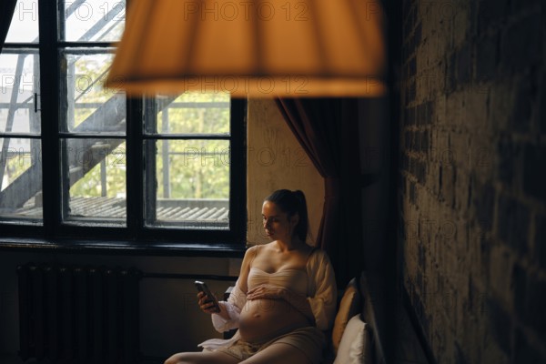 In a cozy room filled with natural light, a pregnant woman sits comfortably on sofa and making