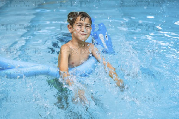 Smiling young boy splashing in a swimming pool, using a blue pool noodle. Joyful summer activity