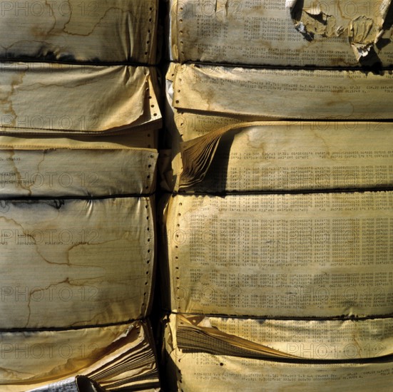 A large stack of compressed paper products is arranged in a recycling area. The paper displays wear