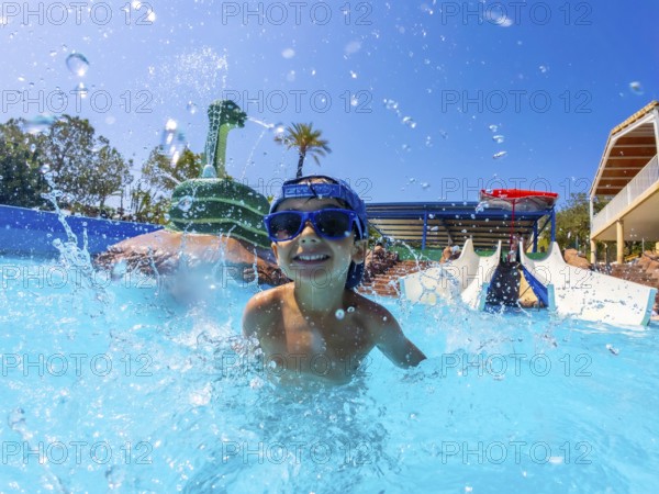 Young boy wearing blue sunglasses, joyfully splashing in a swimming pool at a vibrant water park,