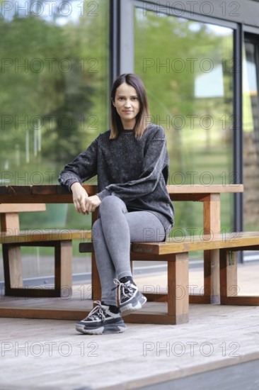 A young woman is seated at a wooden table outdoors, dressed in comfortable athletic wear. She