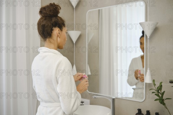 A woman wearing a white robe stands in front of a large mirror in a stylish bathroom. She opens a