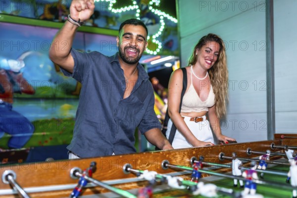 Young man and woman enjoying a foosball game at a funfair, celebrating a goal with excitement and