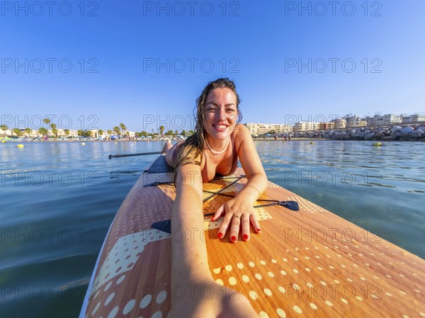 Smiling woman lying on a paddle surfboard, capturing a selfie with a stunning beach and clear blue