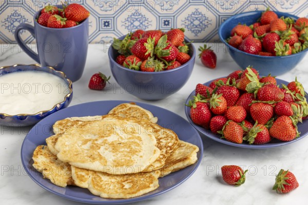 Pancakes on a plate, surrounded by bowls of strawberries and a bowl of yoghurt