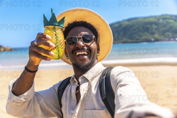 Young man wearing sunglasses and straw hat holding pineapple shaped cocktail while taking selfie on