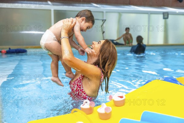 Mother playfully lifting her smiling baby out of the water in an indoor swimming pool during a baby