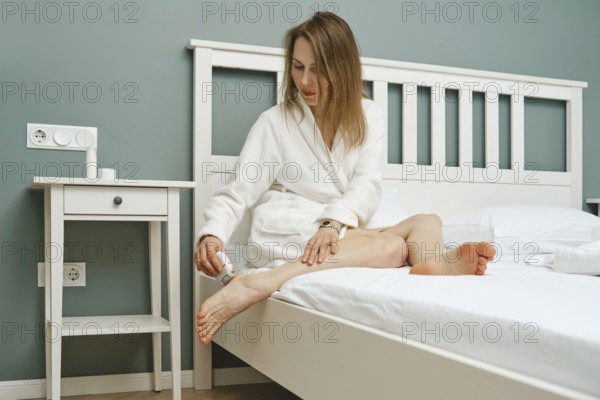 A woman in a white robe sits on the edge of a bed, using electric exfoliating machine on her bare