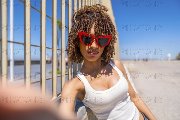 Fashionable young woman with curly hair taking a selfie by the beach, wearing trendy heart shaped
