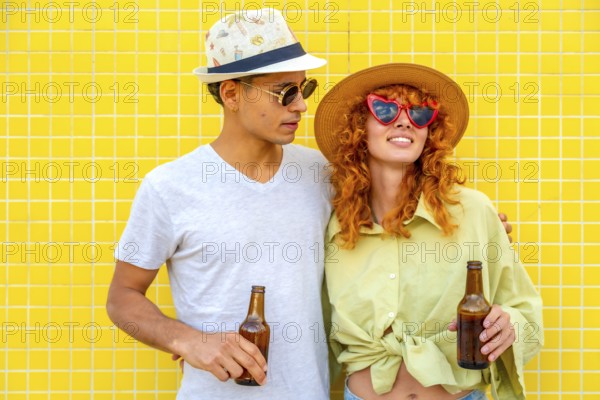 Young couple wearing hats and sunglasses, holding beer bottles and smiling joyfully against a
