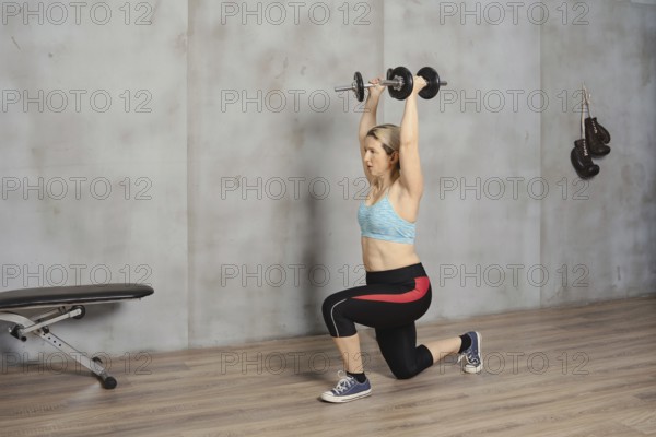 A woman is engaged in a strength training workout, lifting dumbbells overhead while in a lunge