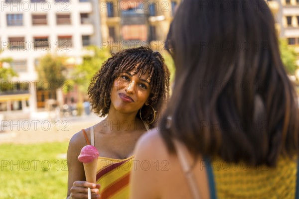 Young woman enjoying a strawberry ice cream cone while talking to her friend in a city park on a