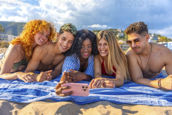 Five cheerful young friends are lying on a beach towel, taking a selfie with a smartphone and