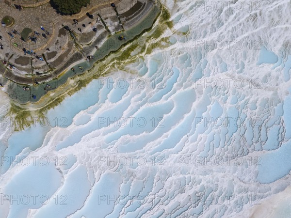 View of limestone terraces with blue water and small figures, surrounded by green vegetation,