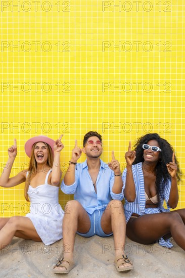 Group of cheerful multi ethnic friends pointing up while sitting on the sand against a vibrant