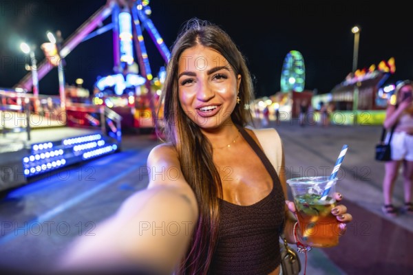 Happy young woman enjoying a vibrant night at an amusement park, taking a selfie while holding a