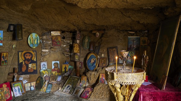 Icon-decorated cave altar with glowing candles, surrounded by religious artworks and paintings,