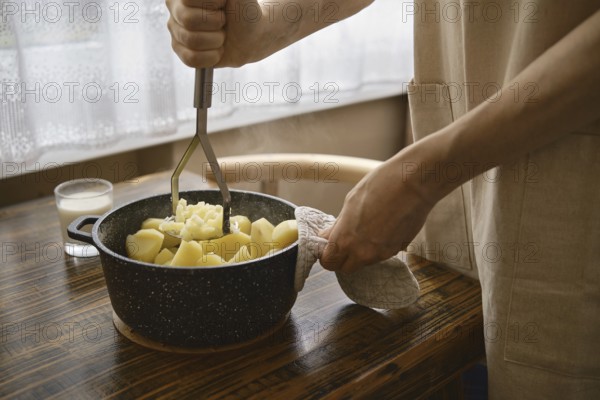 A person is mashing boiled potatoes in a pot. His hand grips a masher while holding a pot with