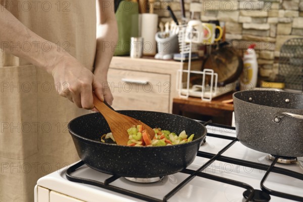 A person stirs a colorful mix of fresh vegetables in a frying pan on a stove in a cozy kitchen.