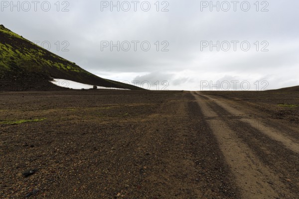 Gravel track, barren volcanic landscape with snowfields, mountain peaks with rain clouds,