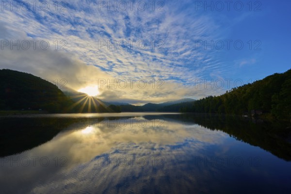 Sunbeams break through clouds and are reflected in a calm lake surrounded by green mountains,