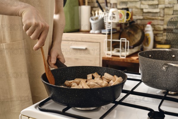 A person stirs diced pork meat in a frying pan on a gas stove. The kitchen is modern, featuring