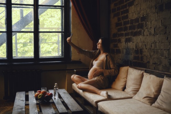 Relaxing at home, a pregnant woman sits on a cozy couch, taking a selfie and posting it in her