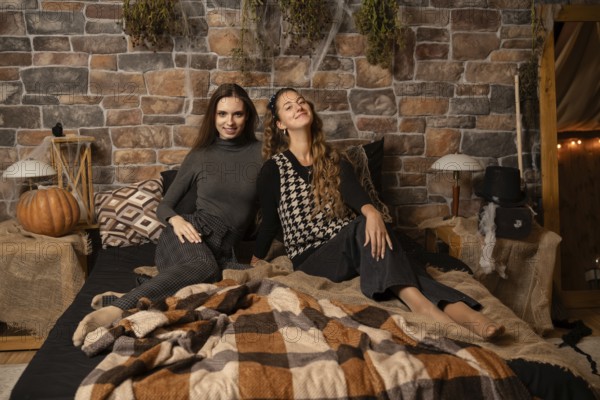 Two women relax together on a bed surrounded by autumn decor, including pumpkins and warm blankets,