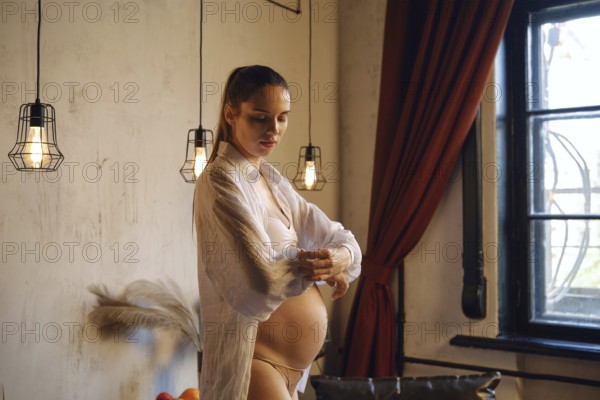 A pregnant woman stands in her elegantly decorated home, adjusting the sleeve of her shirt with a