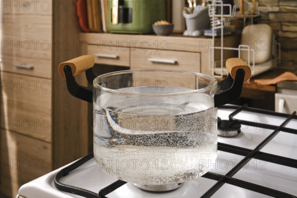 Water is boiling in a clear glass pot placed on a gas stove in a stylish kitchen. The scene