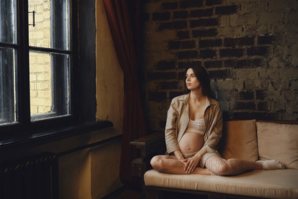 A relaxed pregnant woman sits on a couch in her home, gazing thoughtfully out the window. Soft