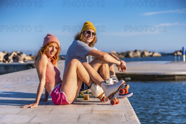 Smiling couple sitting on a sunny pier by the ocean, wearing casual attire and roller skates,