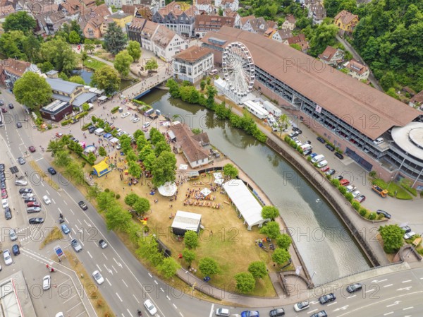 Park with people by a river, flanked by town buildings and a Ferris wheel, with heavy road traffic,