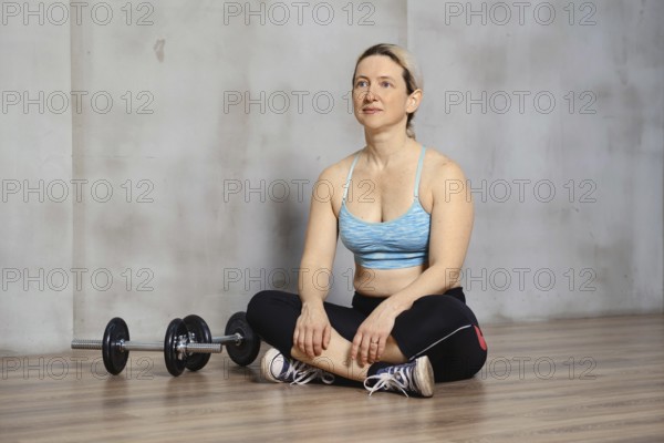 A woman sits cross-legged on a wooden gym floor, dressed in workout attire. Nearby, a pair of