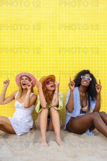 Three diverse women wearing summer clothes and sunglasses are sitting on the sand, pointing upwards