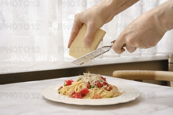 A person grates cheese onto a plate of spaghetti adorned with tomatoes and bacon in a well-lit