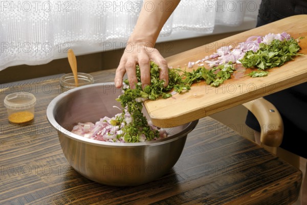 A hand adds chopped cilantro and onions into a silver mixing bowl with minced meat on a wooden