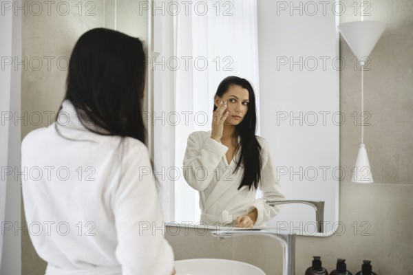 In a bright bathroom, a woman dressed in a white robe applies skincare to her face while looking