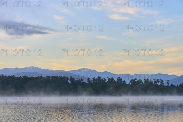 A peaceful lake with a misty surface and mountains in the morning light, Lake Mahinapua, Ruatapu,