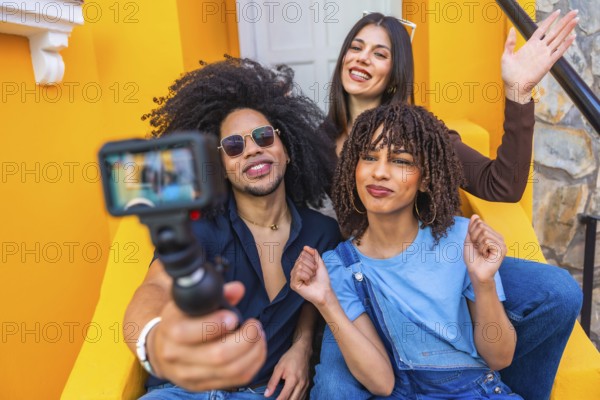 Three happy multi ethnic influencers sitting on yellow steps are filming a vlog with an action