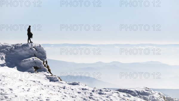 Standing on a rocky ledge, a walker takes in the breathtaking view of snow-covered mountains in