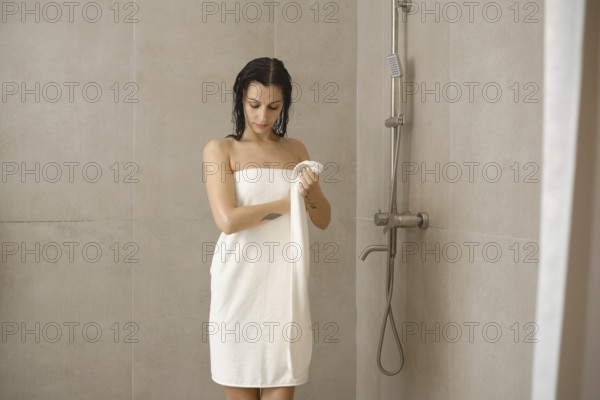 A young woman stands in a modern shower wrapped in a towel, drying her body. The neutral-toned