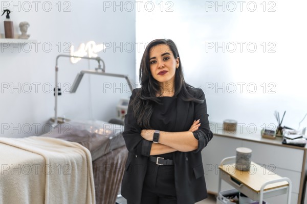 Confident female beautician posing with crossed arms in her modern beauty salon, featuring a laser