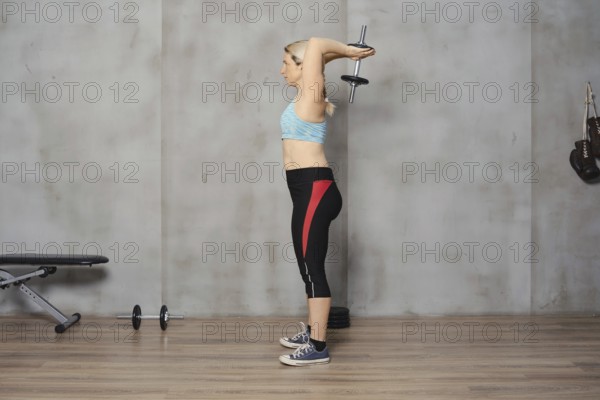 A focused woman in athletic wear is performing a tricep extension with a dumbbell in a