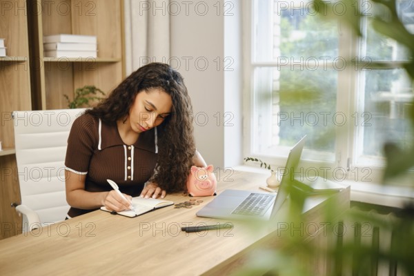 A young woman with curly hair sits at a desk in her bright home office. She writes in a notebook